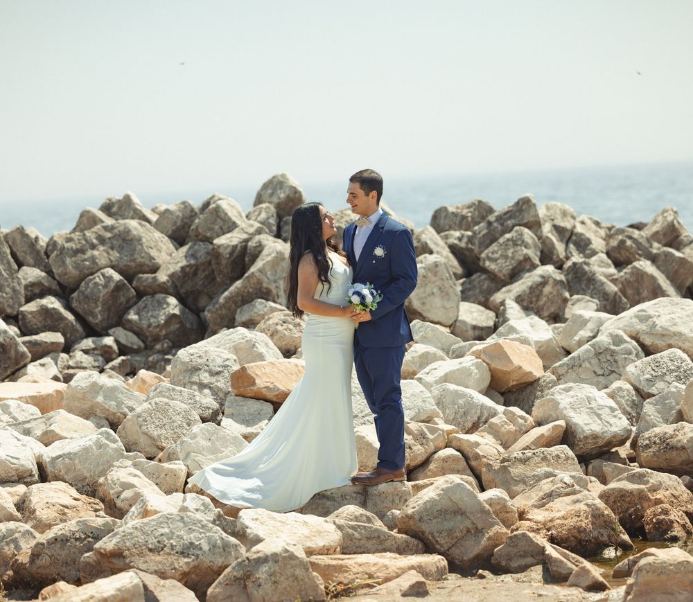 A couple share an intimate moment on rocky coastal terrain in formal wedding attire.