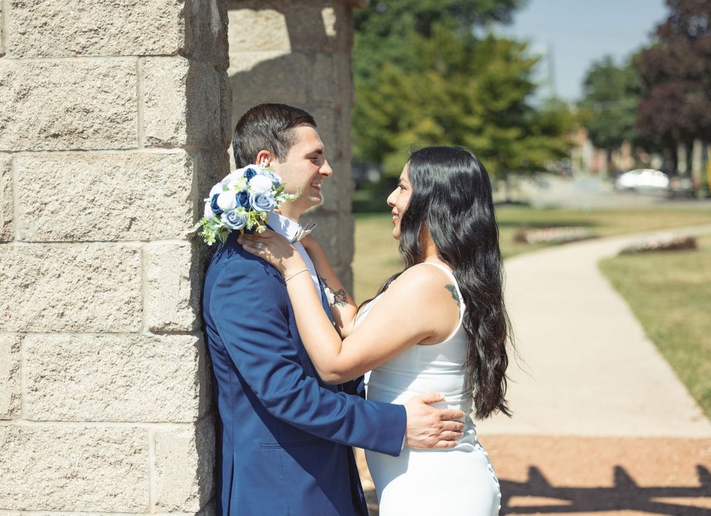 A couple in wedding attire embrace outdoors next to a stone wall on a sunny day.