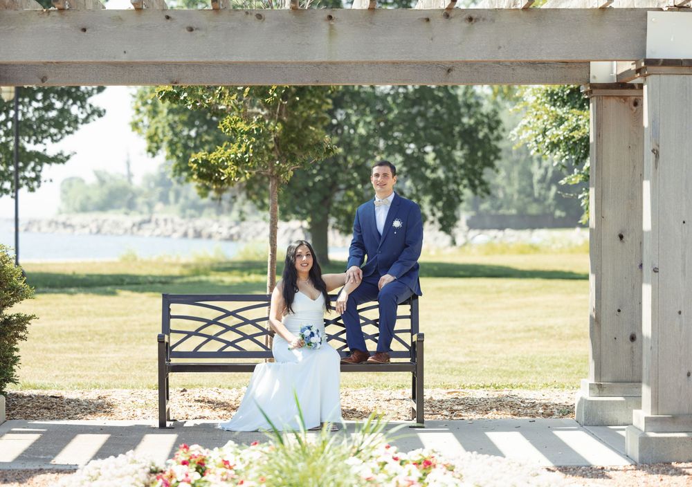 Couple standing in white gazebo on a sunny day next to a lake with trees and landscaping in the background.