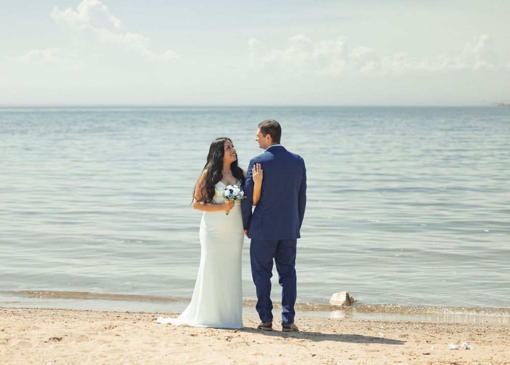 Newlywed couple walking along a serene beach shoreline with gentle waves in the background.