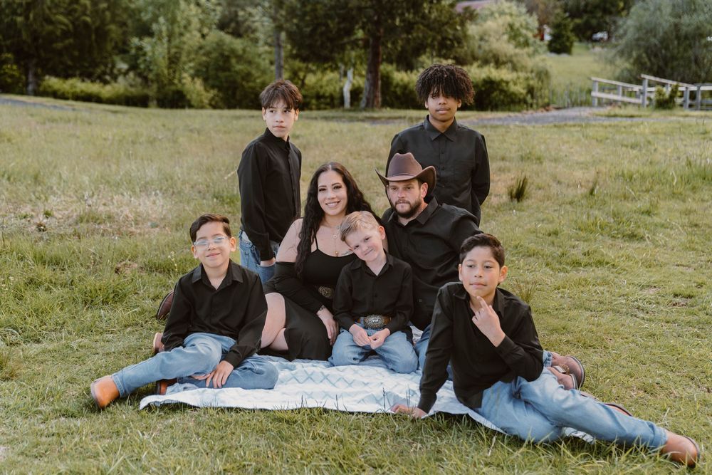 Family of seven sitting together on a blanket in a green meadow.