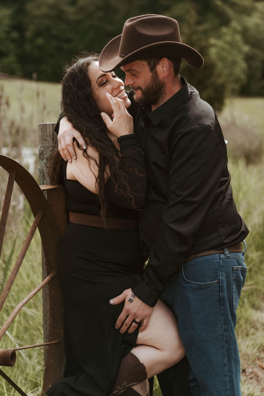 Couple embracing near a rustic wagon wheel in a countryside setting.