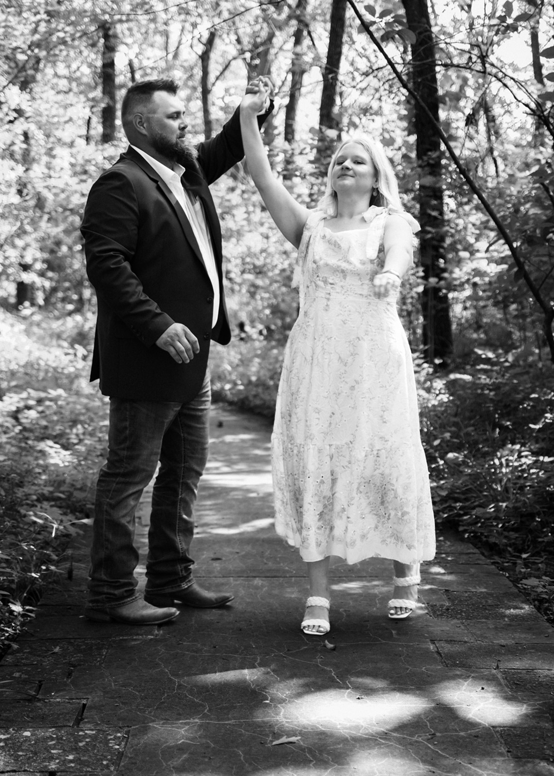 Man twirling his bride to be around on concrete pathway through Powell Gardens. White floral dress flowing as she twirls