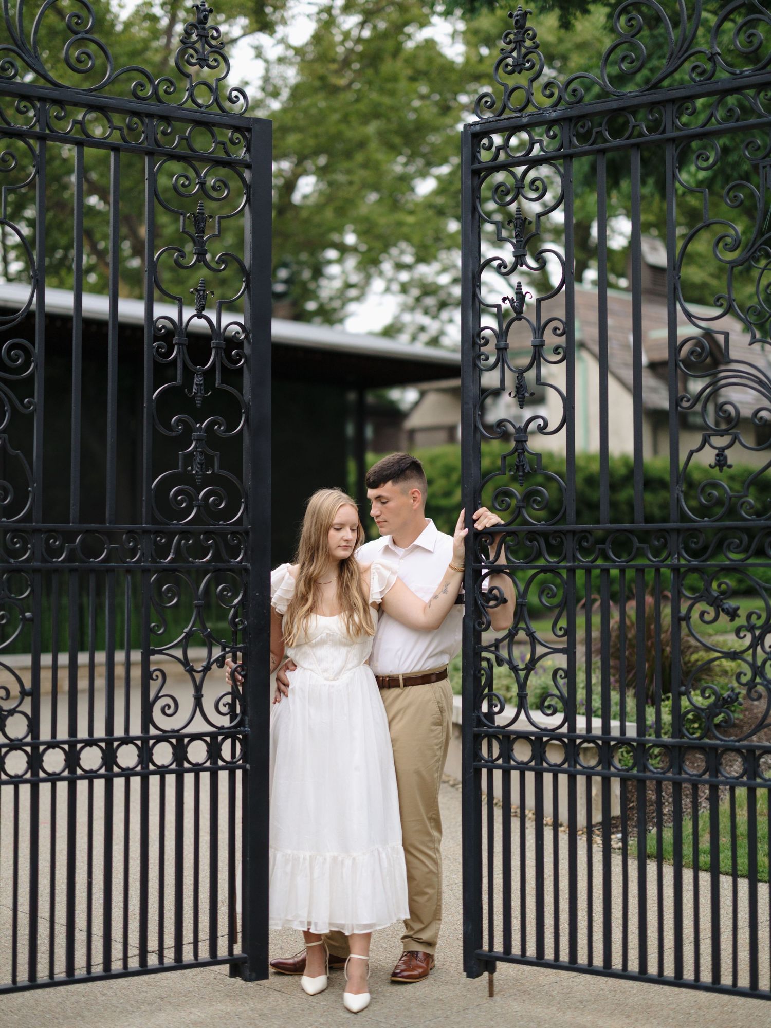 A couple stands together at ornate wrought iron gates in a garden setting, with the person wearing a flowing white dress.
