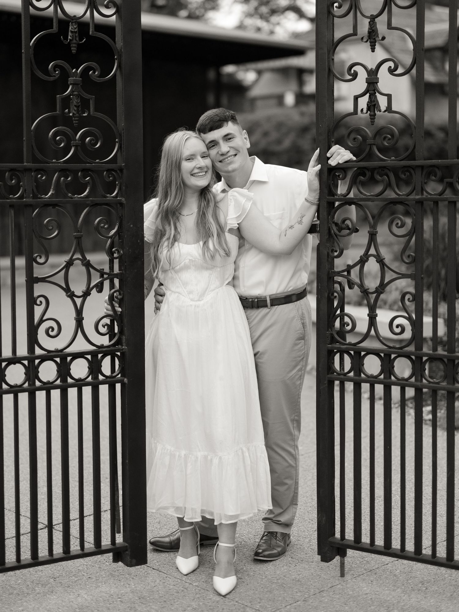 Black and white romantic portrait of a couple standing between ornate iron gates in formal attire.