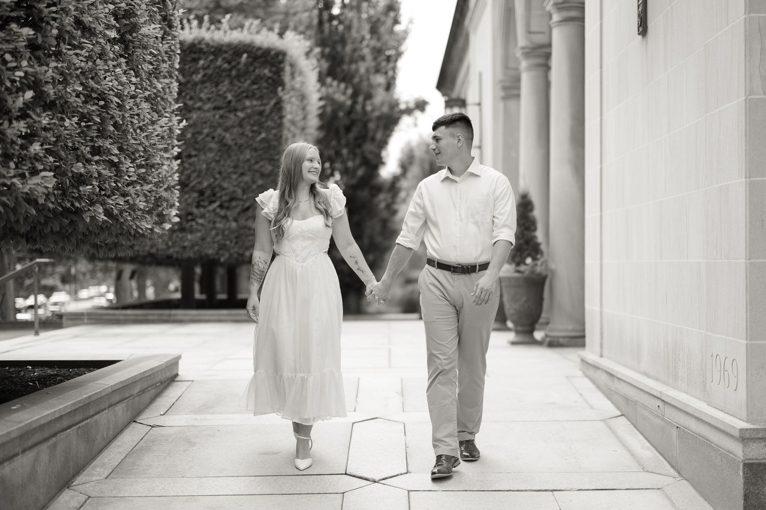 A couple in black and white walking hand in hand along a columned walkway in formal attire.