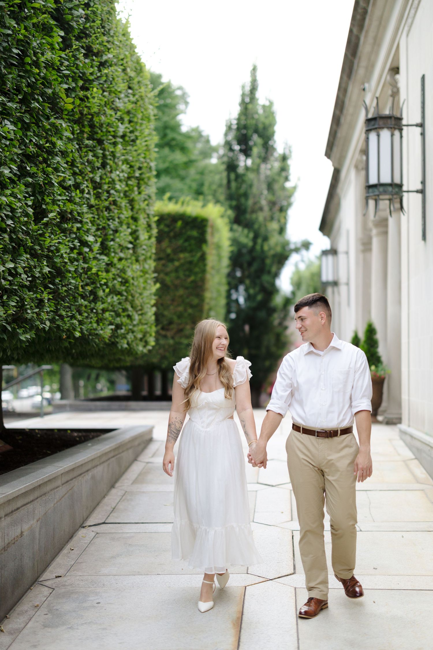 A couple walks hand in hand along an elegant walkway with manicured hedges and classical architecture.