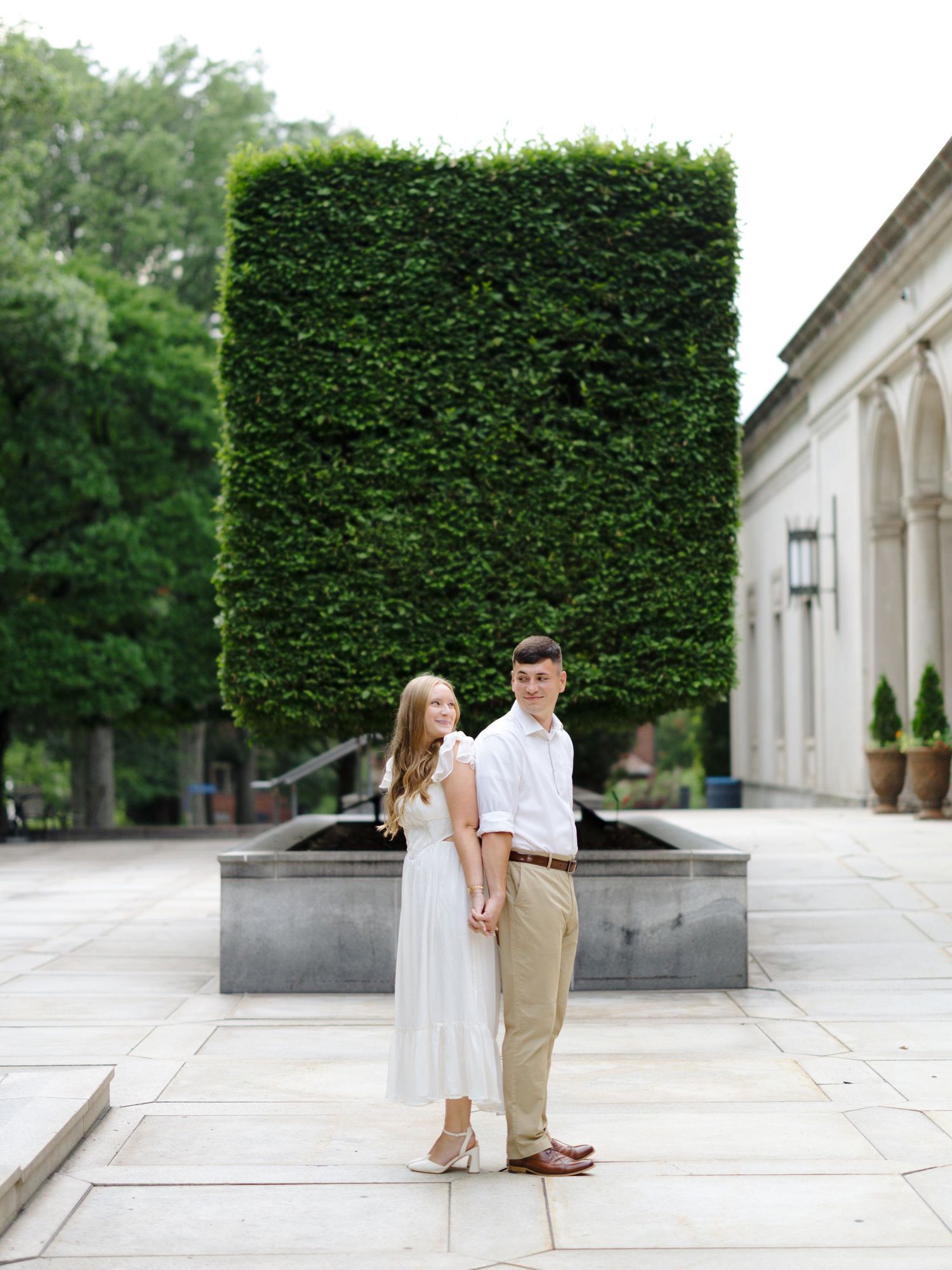 Couple stands together on elegant stone patio with tall manicured hedge and white columns in background.