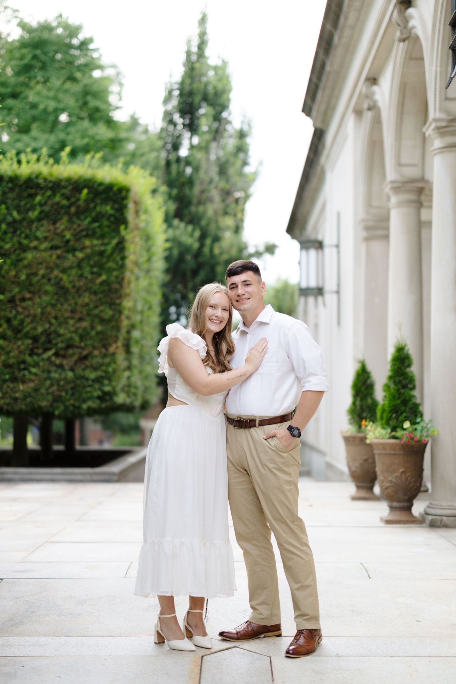 A couple in white and khaki embrace on an elegant stone porch with manicured hedges and classical columns.