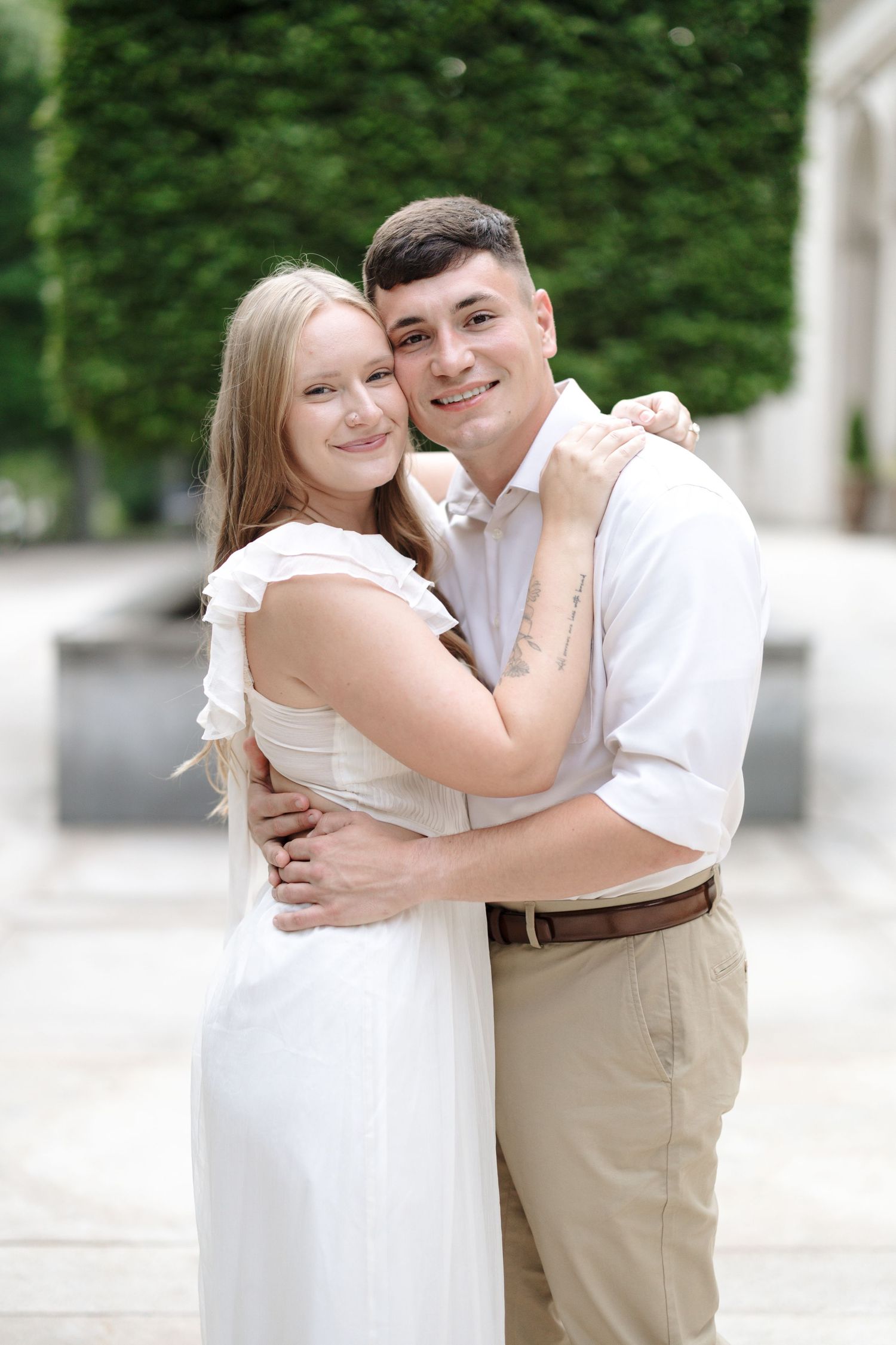 A romantic couple in white and khaki embraces while posing outdoors against a leafy green background.
