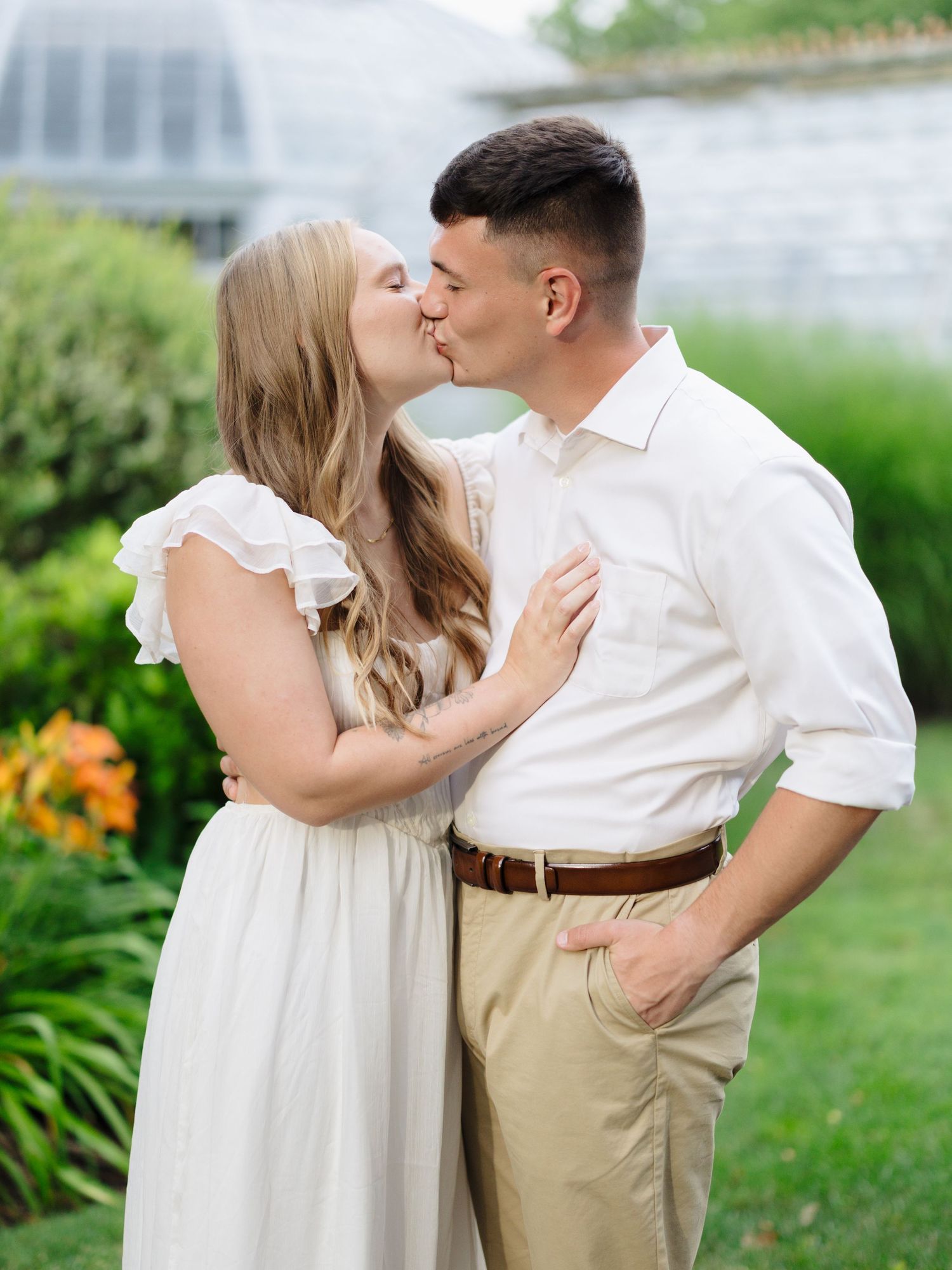 A romantic couple shares a tender kiss outdoors in a garden setting, she in white and he in khaki pants.