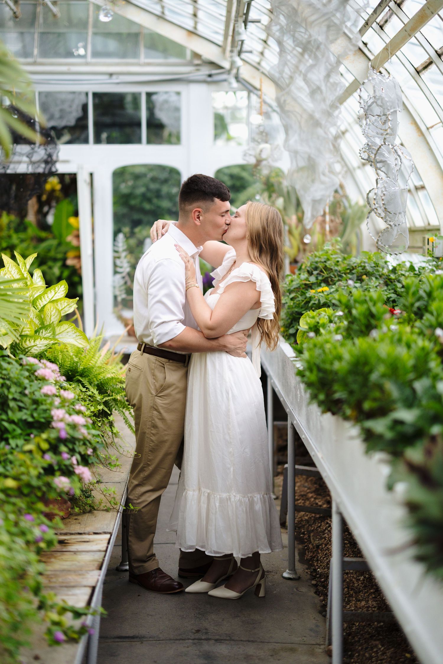 A couple shares an intimate moment in a sunlit greenhouse filled with lush plants and greenery.