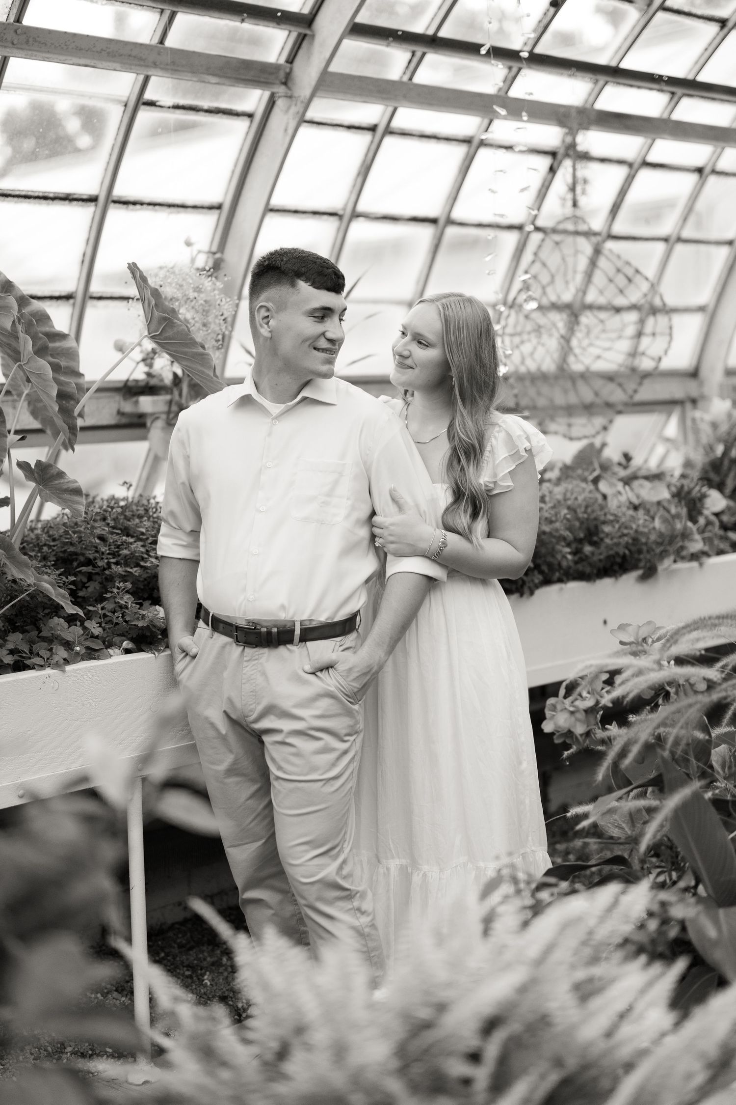 A romantic couple shares tender moments in a greenhouse filled with lush plants and flowers.