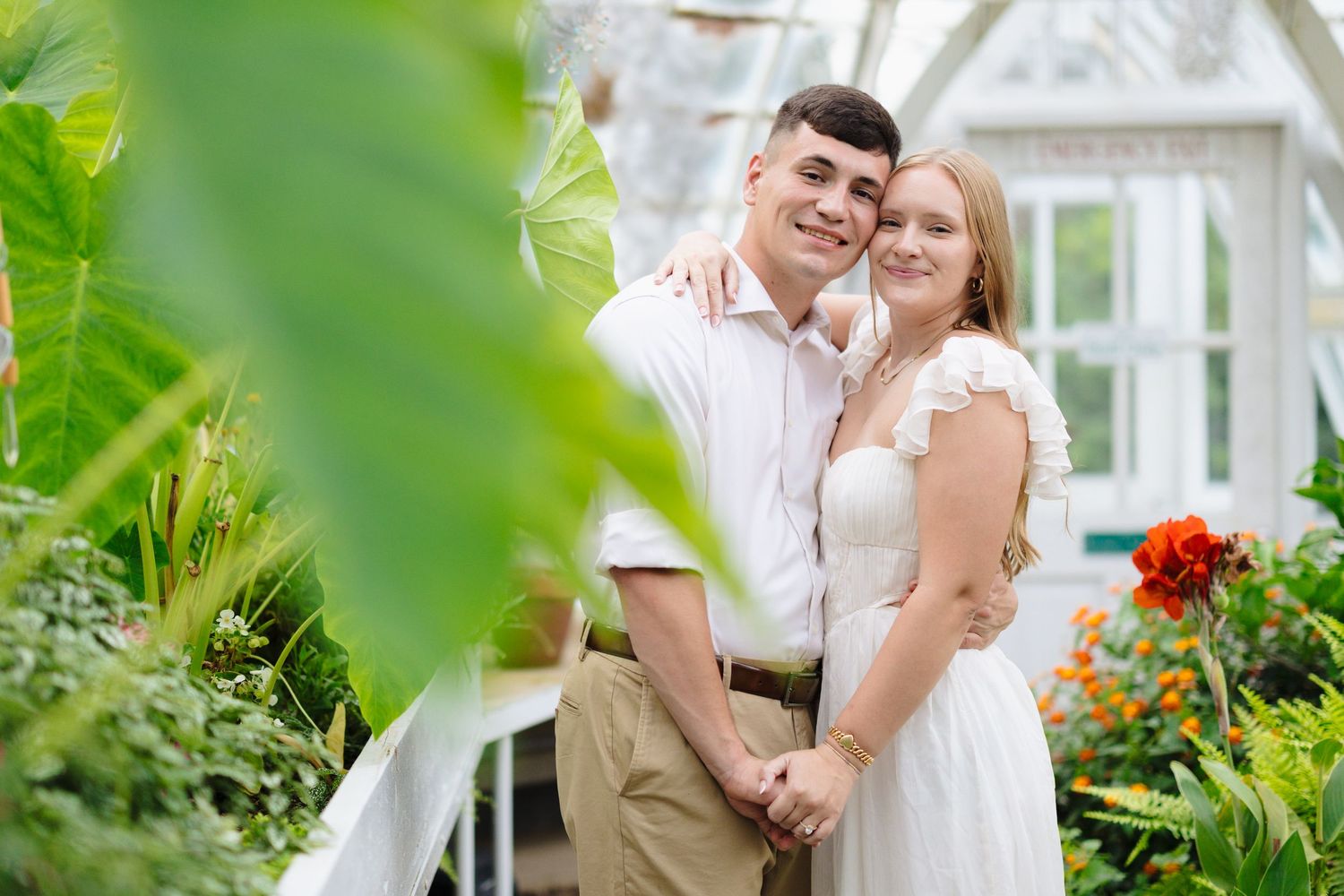 A couple poses together in a garden setting with vibrant greenery and flowers near a white structure.