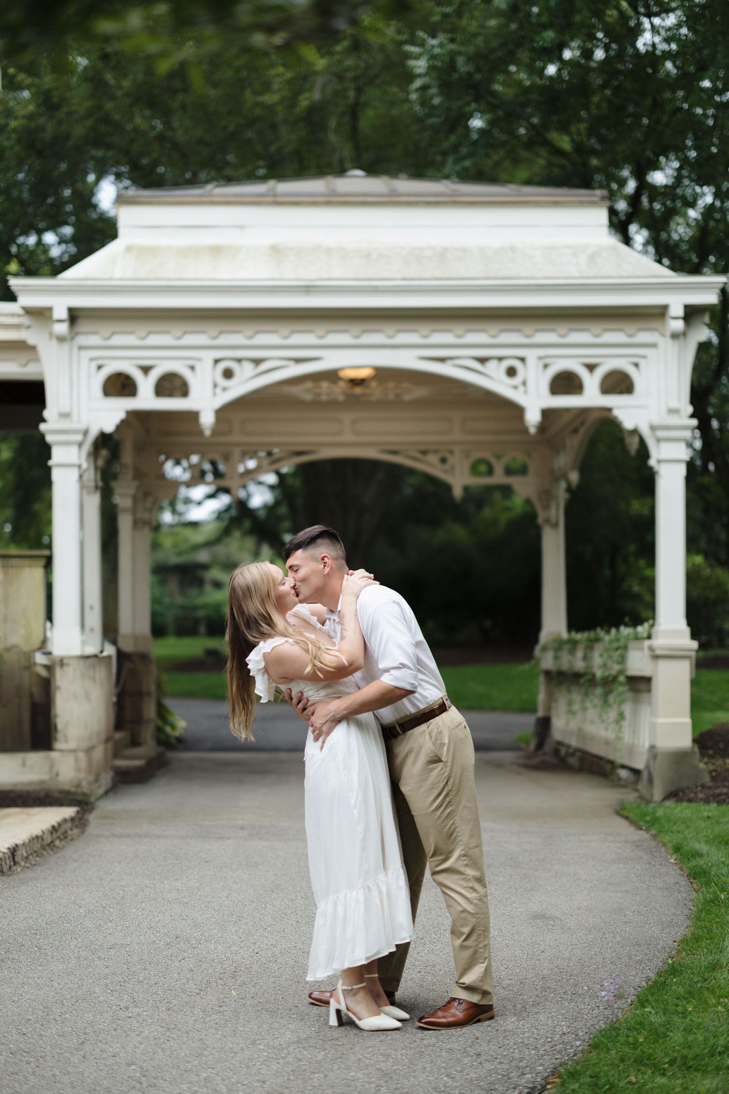 A romantic couple shares tender kisses under an ornate white pergola walkway in a garden setting.