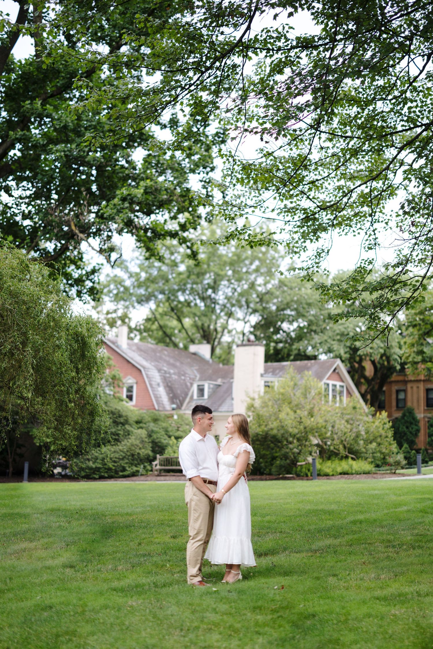 A couple embraces on a lush green lawn in front of a traditional brick home surrounded by trees.