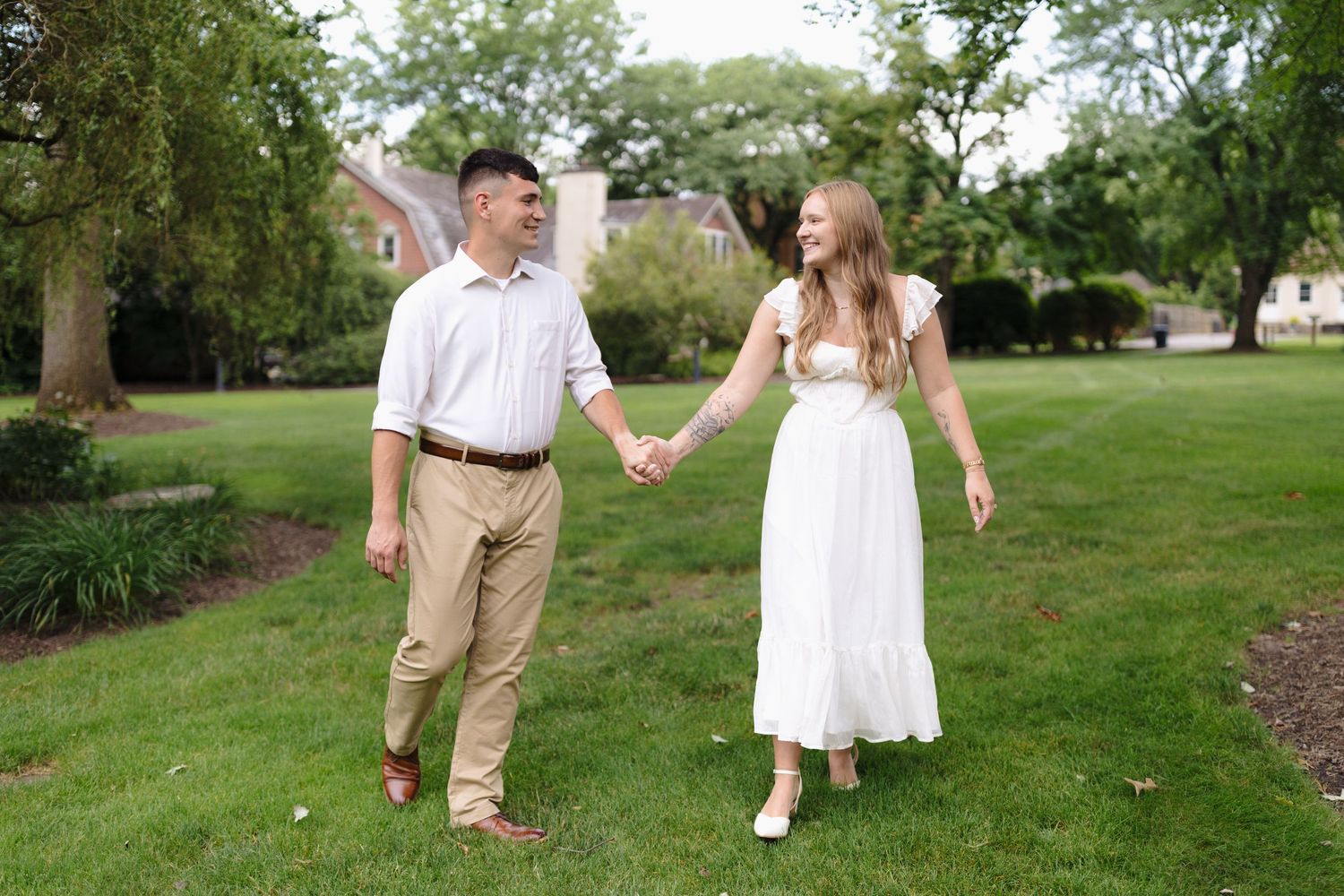 A romantic couple walks hand in hand on a lush green lawn with trees and gardens in the background.