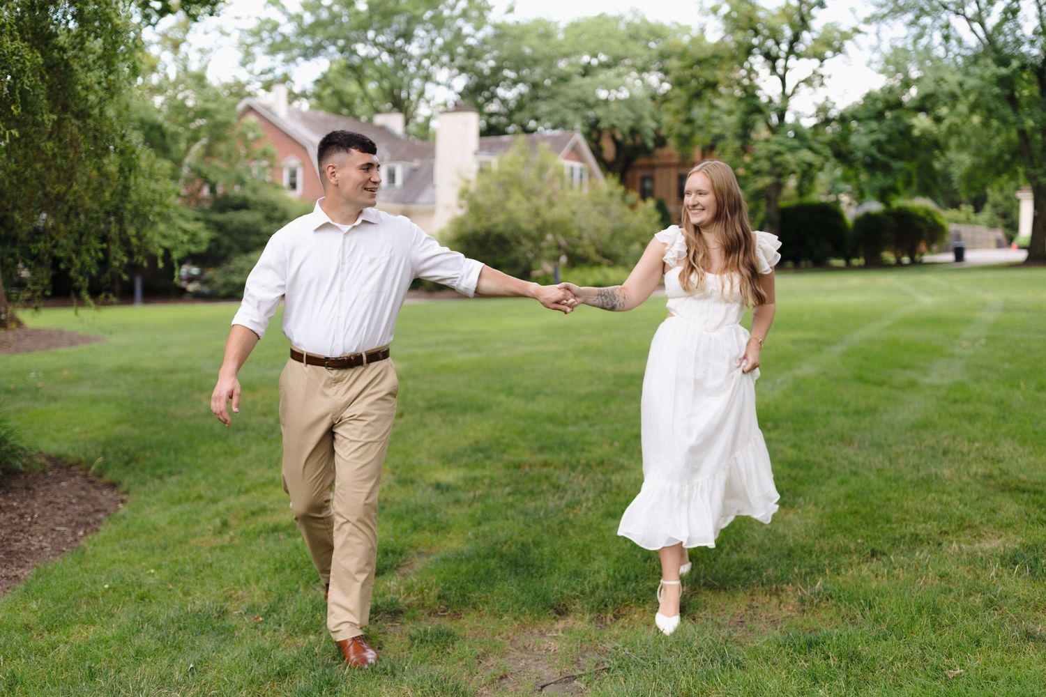 A couple in coordinated light summer outfits walks hand in hand on a lush green lawn near blooming gardens.