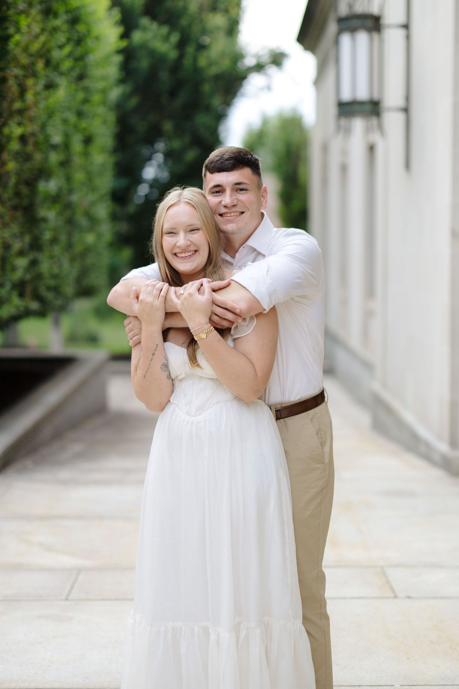 A romantic outdoor portrait showing a couple embracing, with her in a white dress and him in khakis and white shirt.