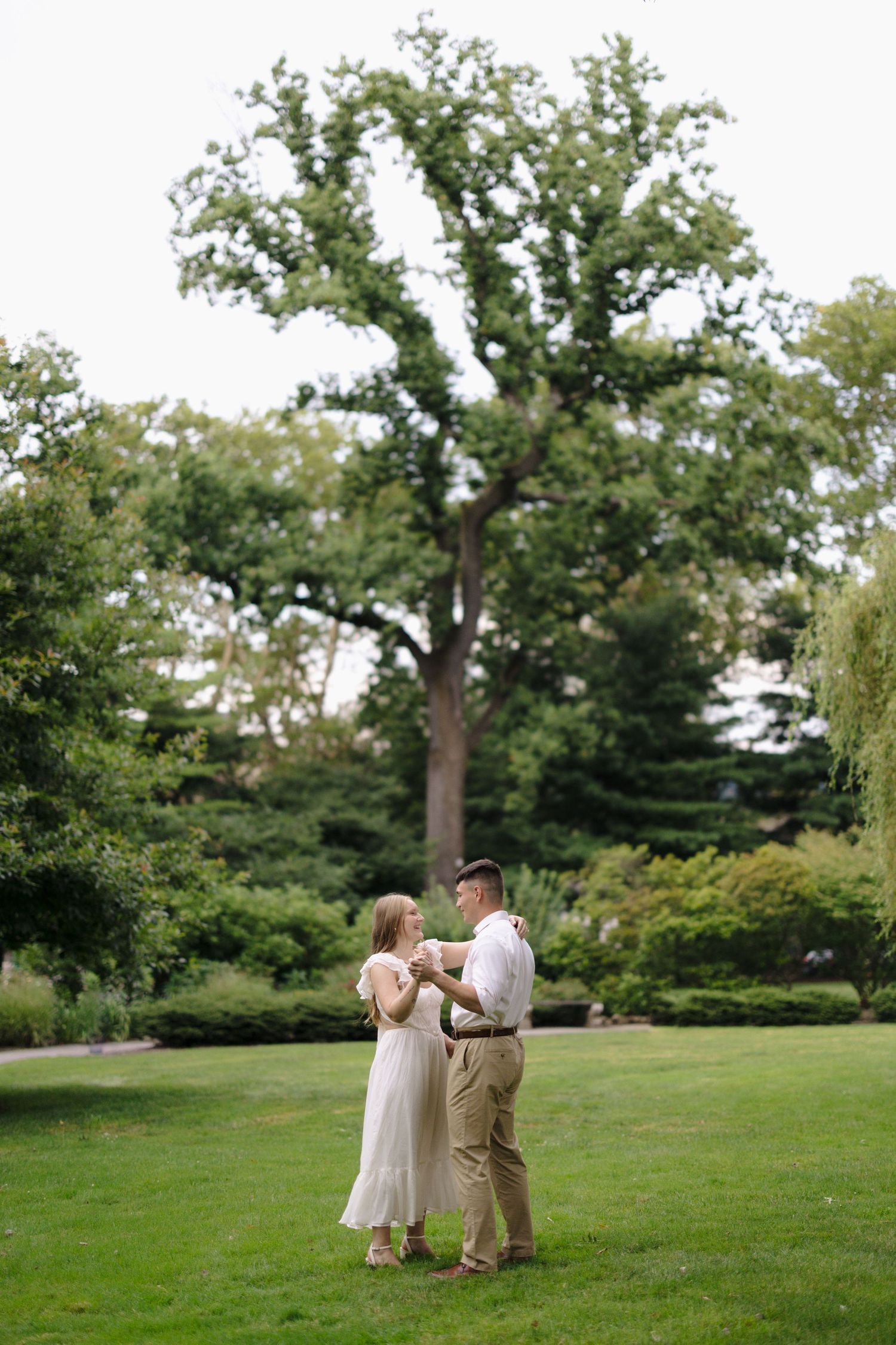 A couple shares romantic moments on a lush green lawn with mature trees in the background.