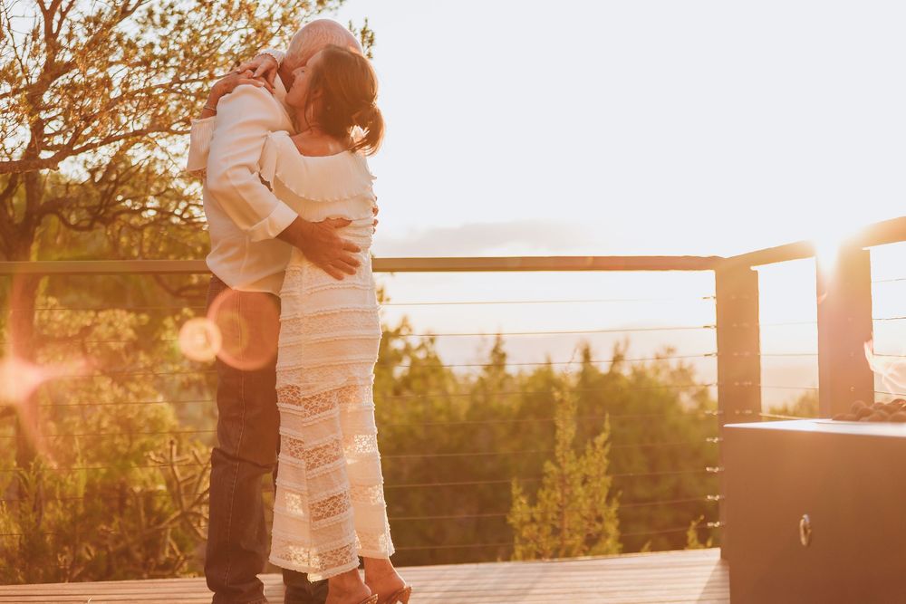 A couple shares a romantic embrace outdoors at golden hour with sunlit trees in the background.