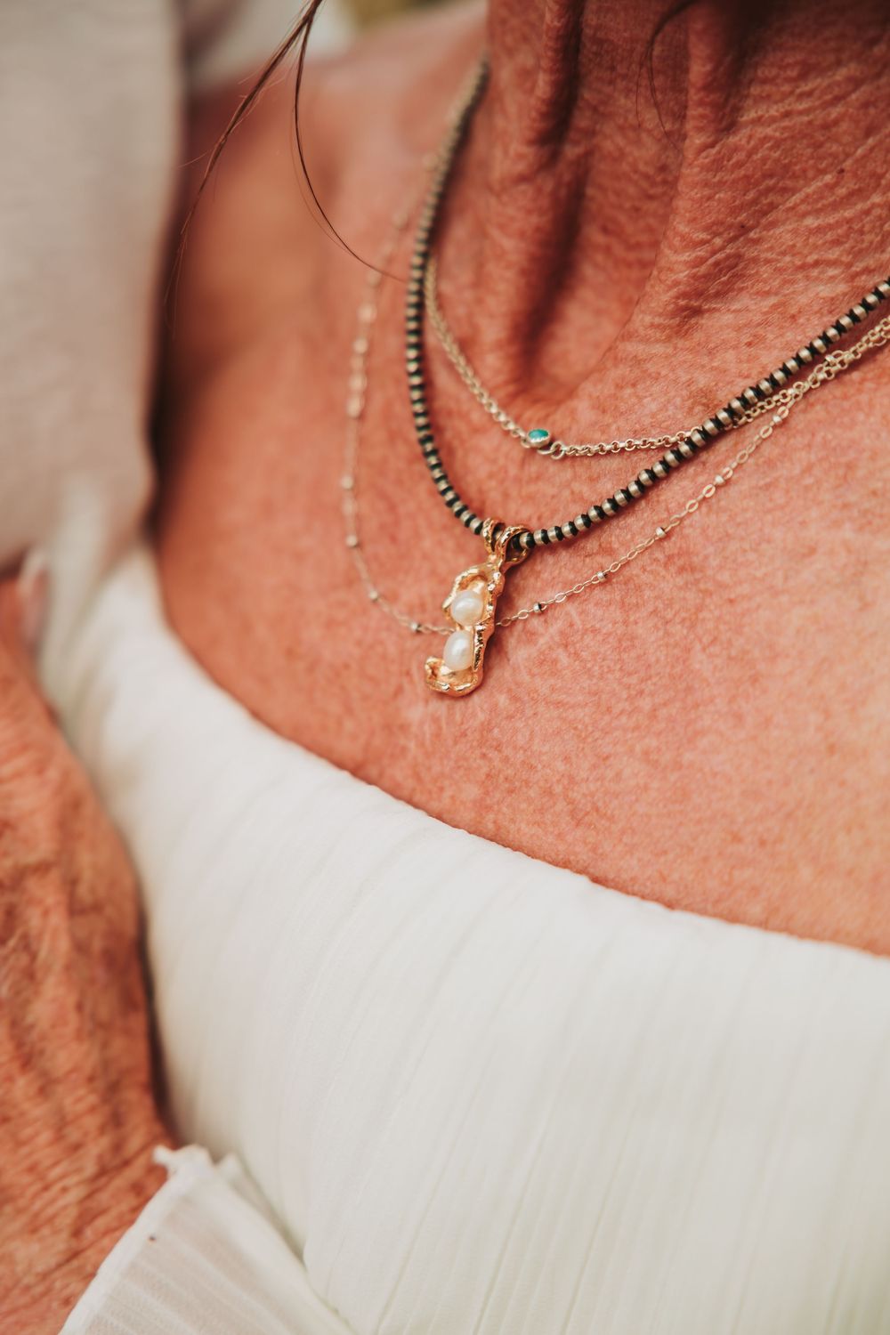 Close-up of layered silver necklaces with a delicate pendant against white fabric and sunlit skin.