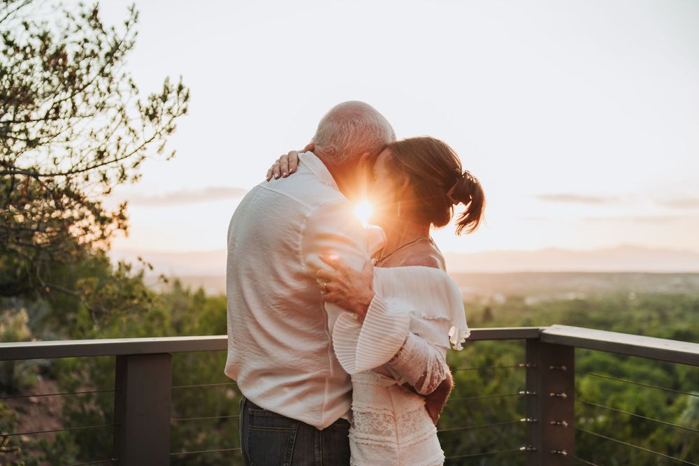 Two people share a romantic sunset embrace on a scenic overlook balcony surrounded by lush greenery.