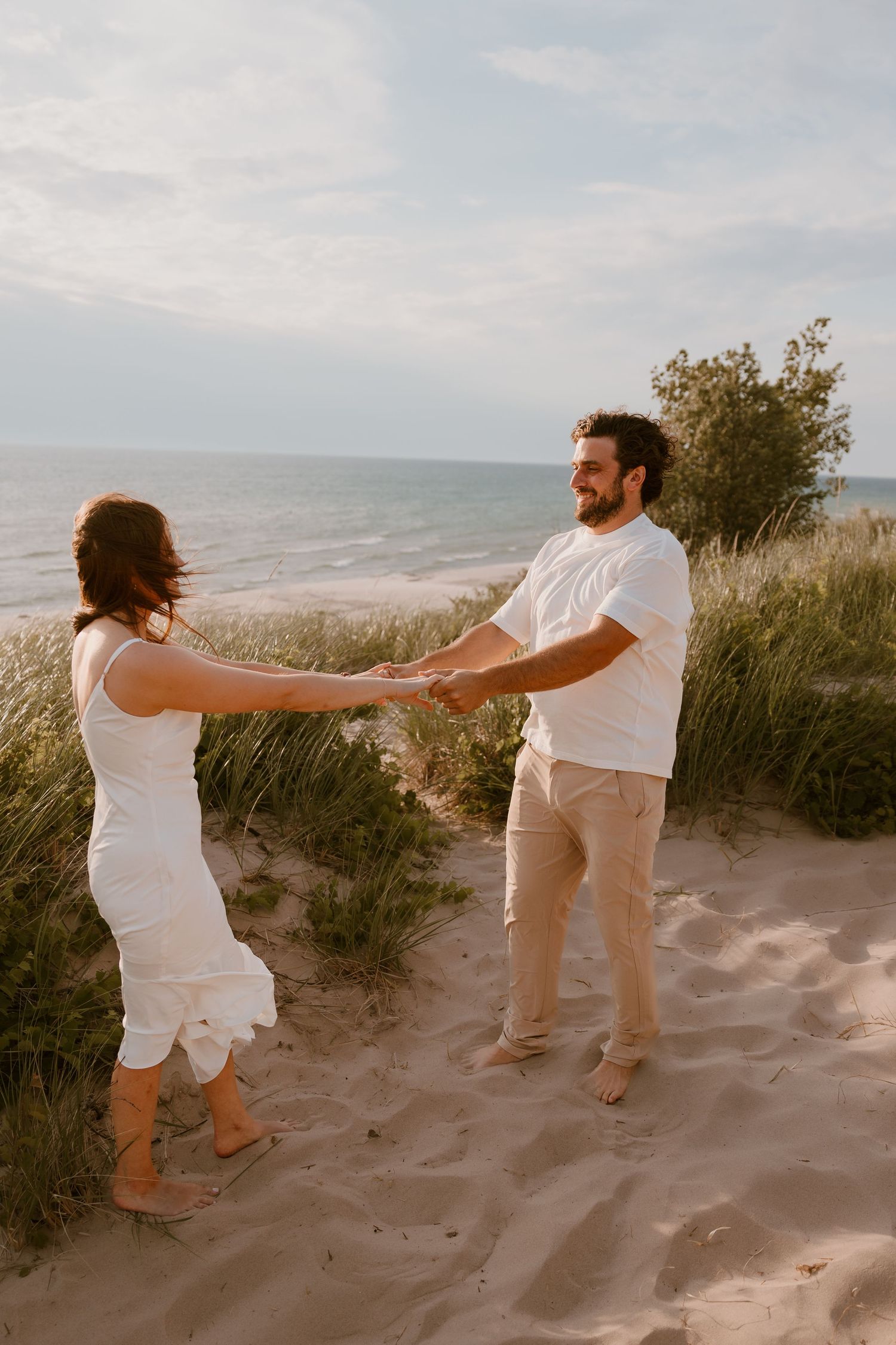 A couple in white outfits holds hands playfully on sand dunes at sunset near beach grass and ocean. Metro Detroit Wedding Photography