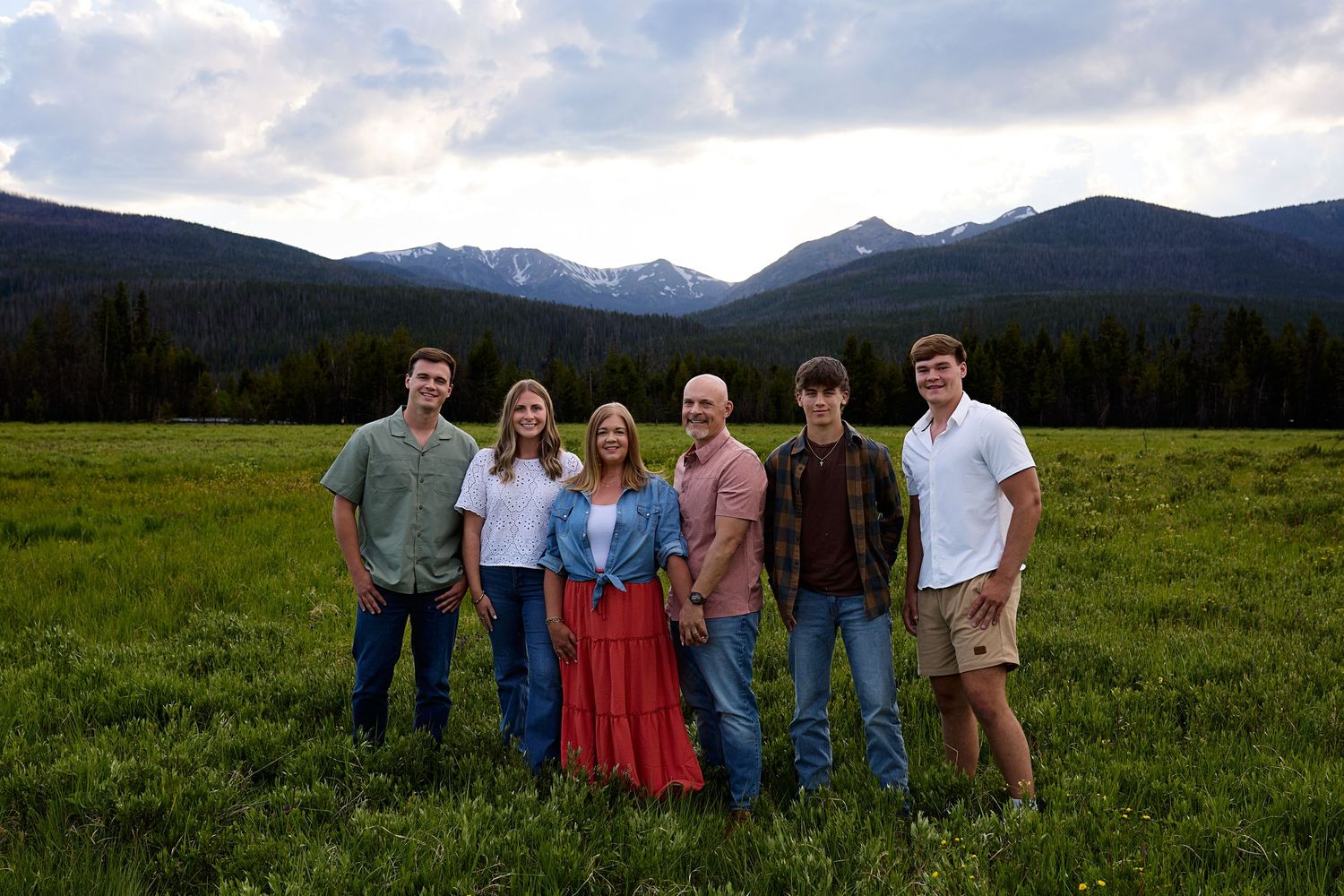 A group stands together in a scenic mountain meadow with snow-capped peaks and evergreen forest in the background.
