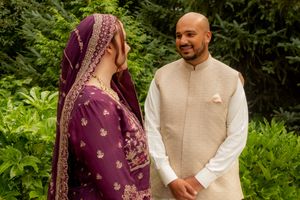 Couple in traditional South Asian wedding attire, bride in purple dupatta and groom in beige sherwani, posing outdoors.