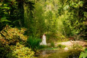 Bride in white dress standing by a serene forest stream surrounded by lush green trees and vibrant foliage.