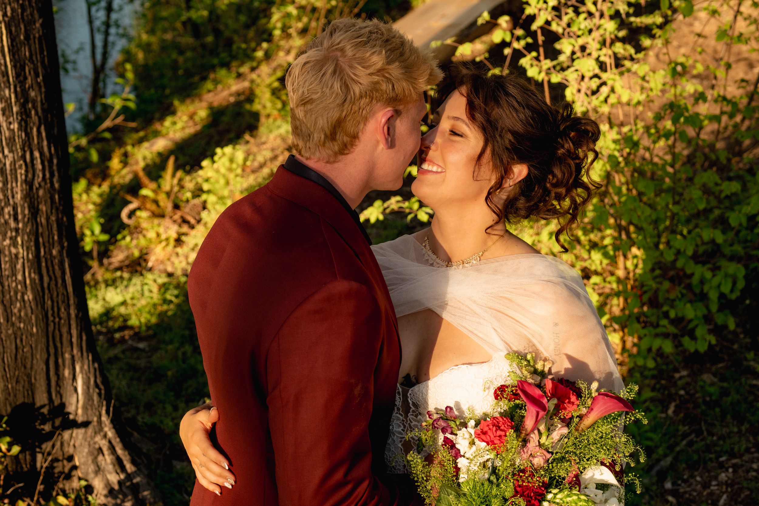 A couple shares a romantic moment outdoors in autumn, bride holding red bouquet in white gown, groom in burgundy suit.