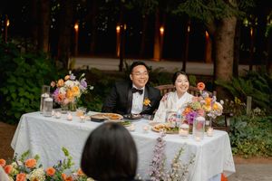 Couple in wedding attire smiling at floral-decorated sweetheart table during outdoor evening reception.