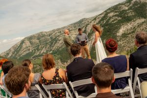 Outdoor wedding ceremony with mountain backdrop, guests seated in white chairs watching couple exchange vows.