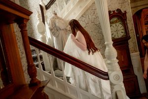 Bride in white gown descending ornate wooden staircase beside grandfather clock in elegant Victorian home.