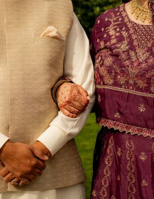 Indian couple in traditional wedding attire, bride with mehndi henna hands holding groom in cream sherwani and purple embroidered lehenga.