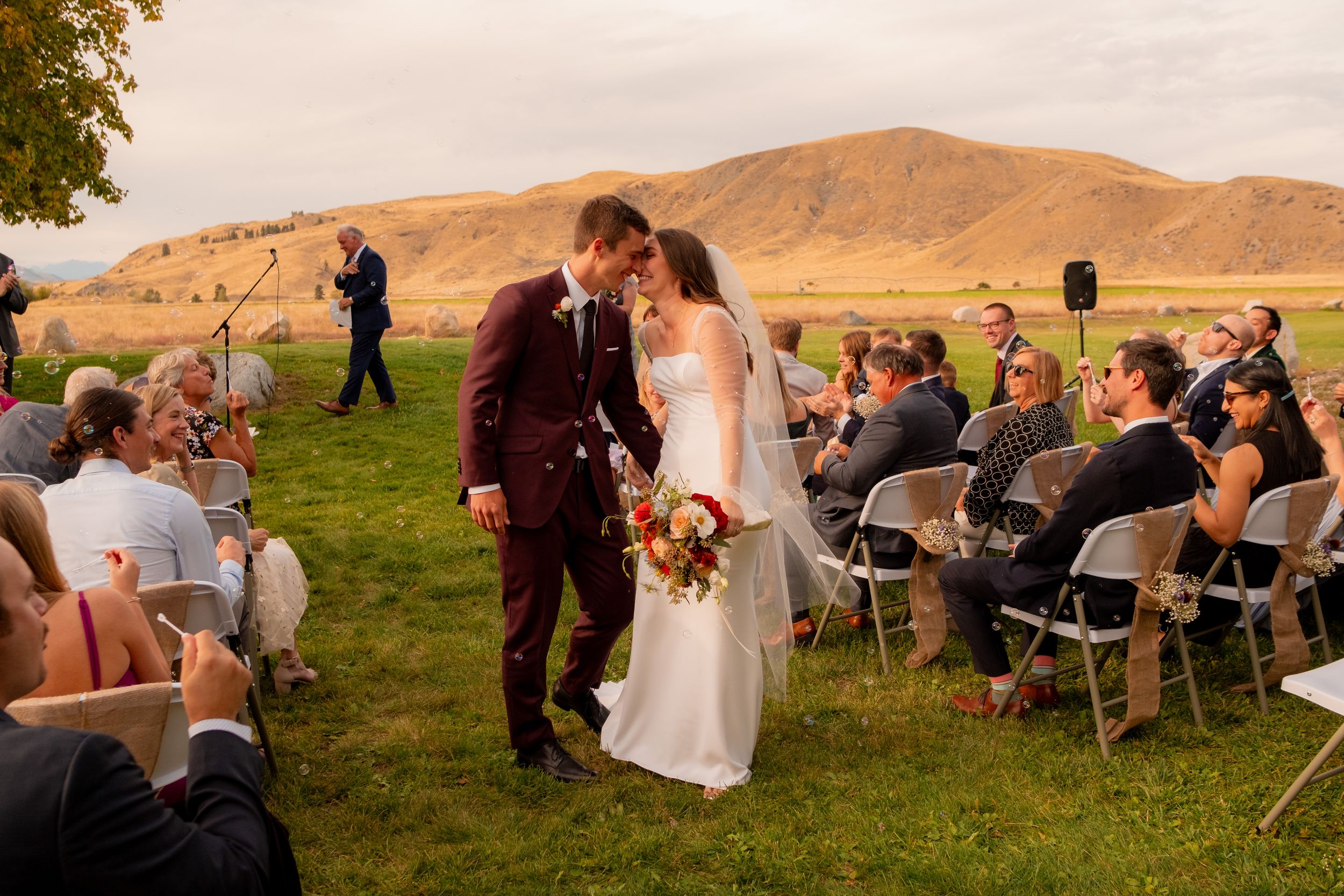 Newlyweds share first kiss at outdoor ceremony with mountain backdrop and seated guests.