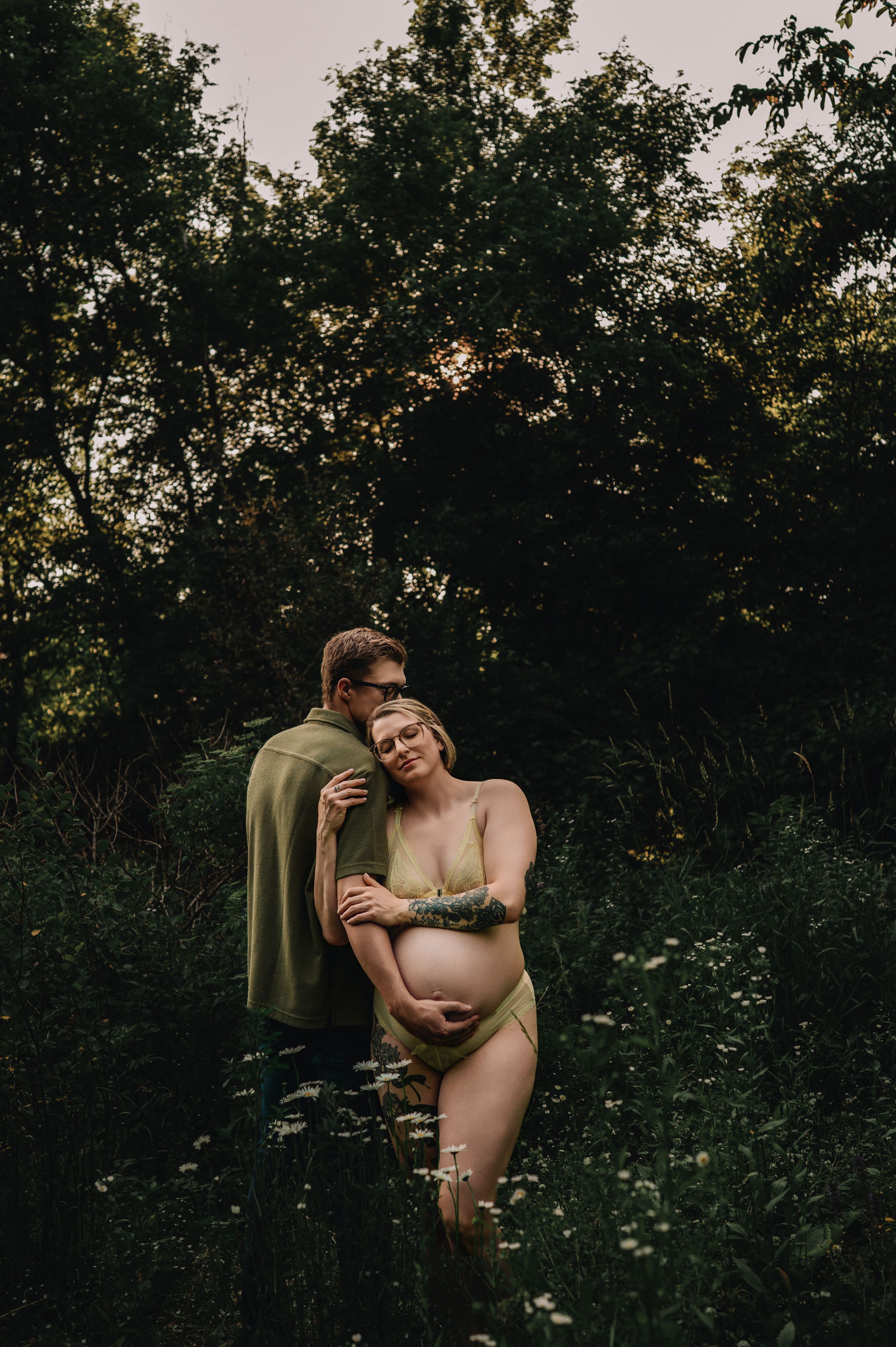 Intimate outdoor maternity photoshoot in a natural setting with greenery and soft evening light.