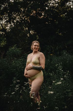 A pregnant person wearing beige swimwear poses outdoors in a nature setting during a maternity photoshoot.