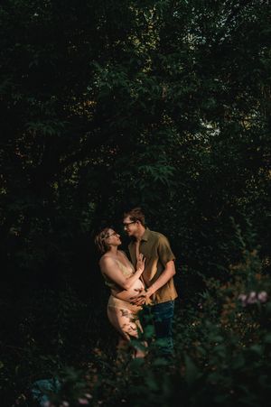 A romantic couple embraces intimately in a dark forest setting surrounded by lush greenery and foliage.