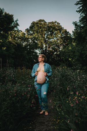 Expecting mother in blue jacket and jeans stands on nature trail at sunset among wildflowers and trees.