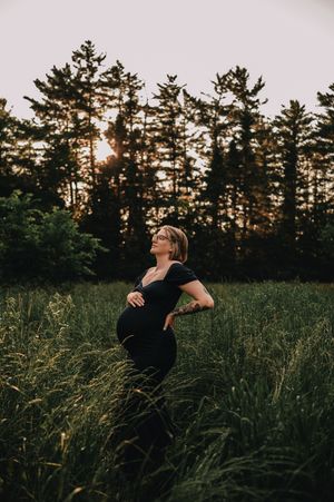 Silhouette of person in black shirt standing in tall grass field at sunset with pine trees in background.