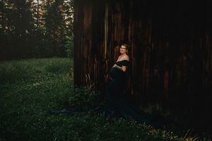 A figure in a black gown stands against a rustic wooden wall in a forest setting at dusk.