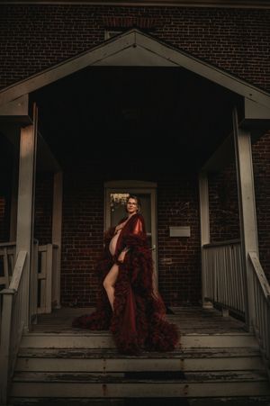 Dramatic photo of figure in flowing red gown on porch steps of brick building at dusk.
