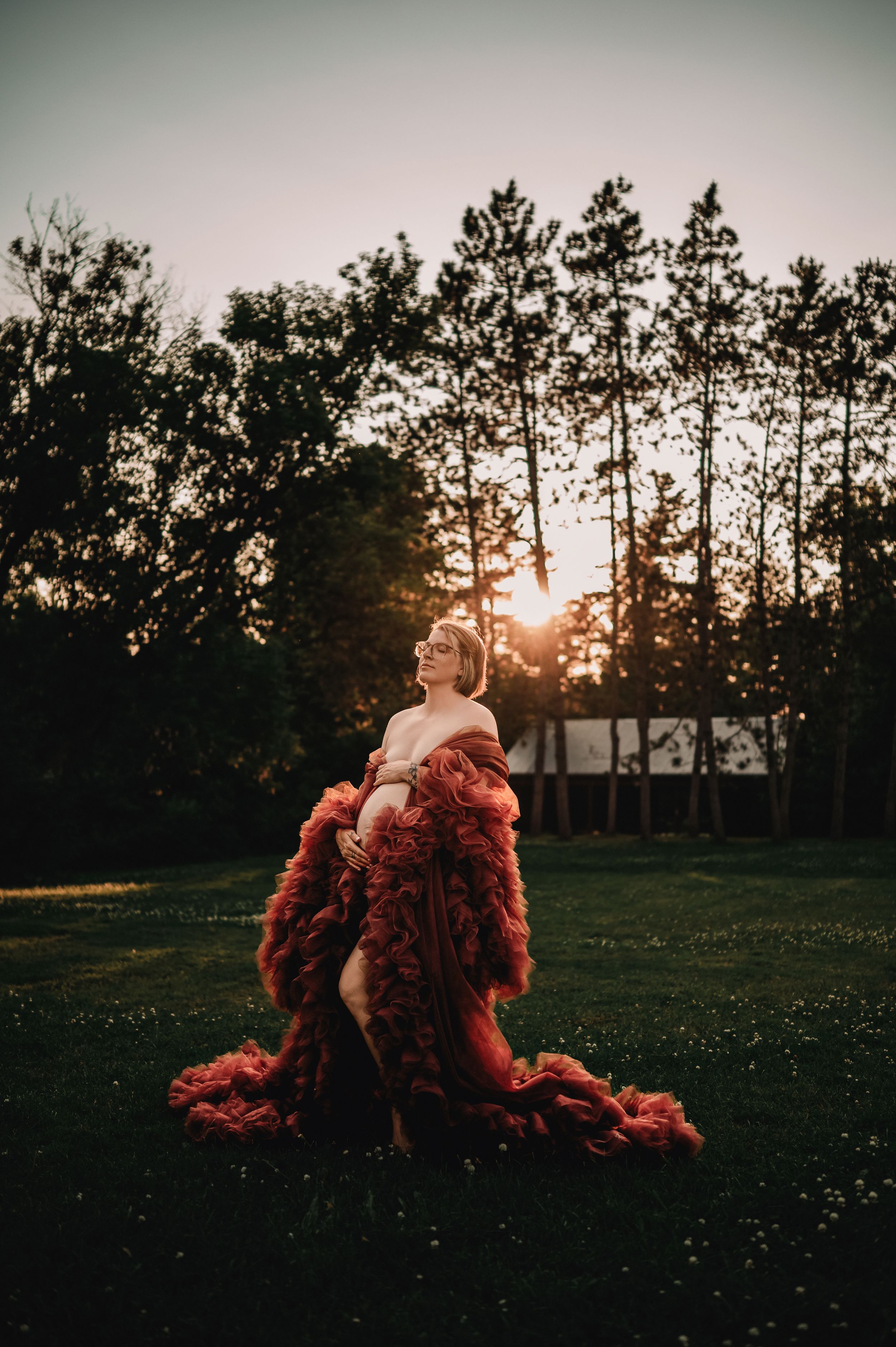 A dramatic silhouette of a person in a flowing red dress against a sunset through pine trees in a grassy field.