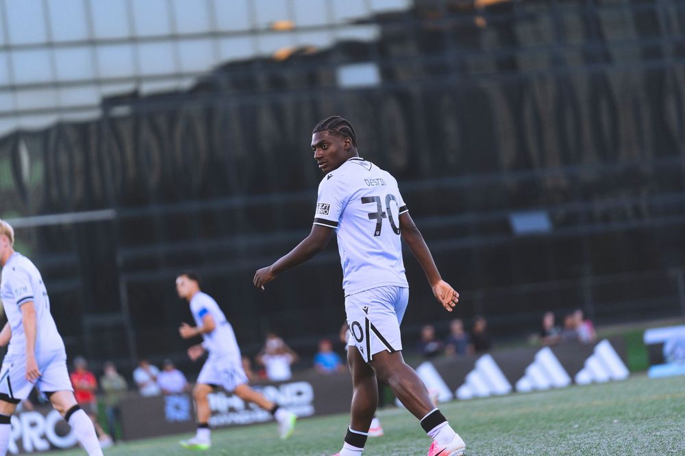 A soccer player walks on the field with other players and fans in the background