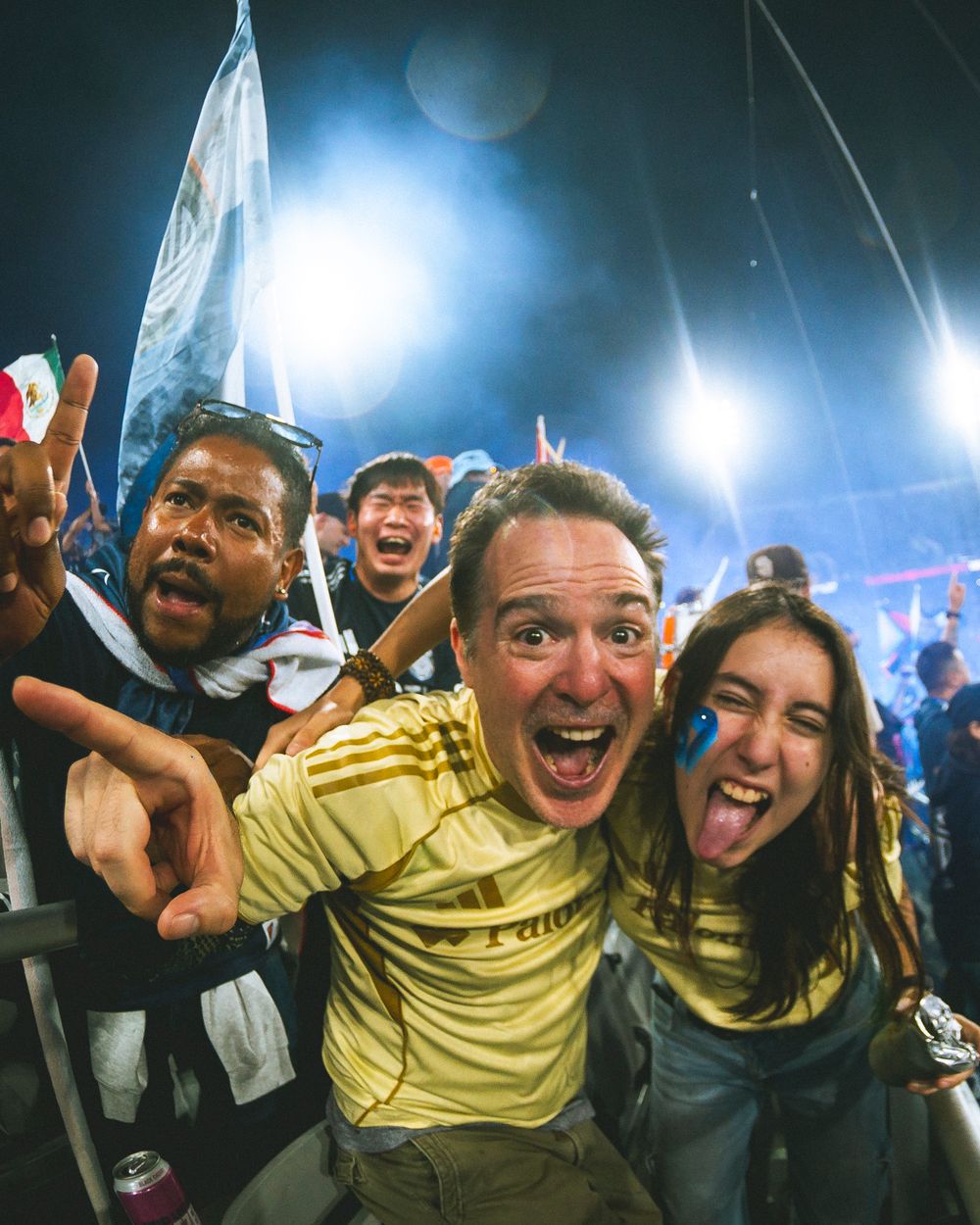 Two fans celebrate a goal, smiling and pointing at the camera with blue smoke and bright lights in the background