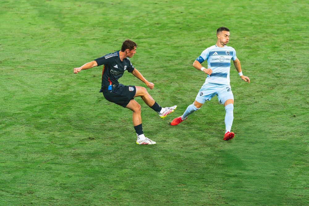 Two men play on a soccer field one kicking the ball and the other chasing it.