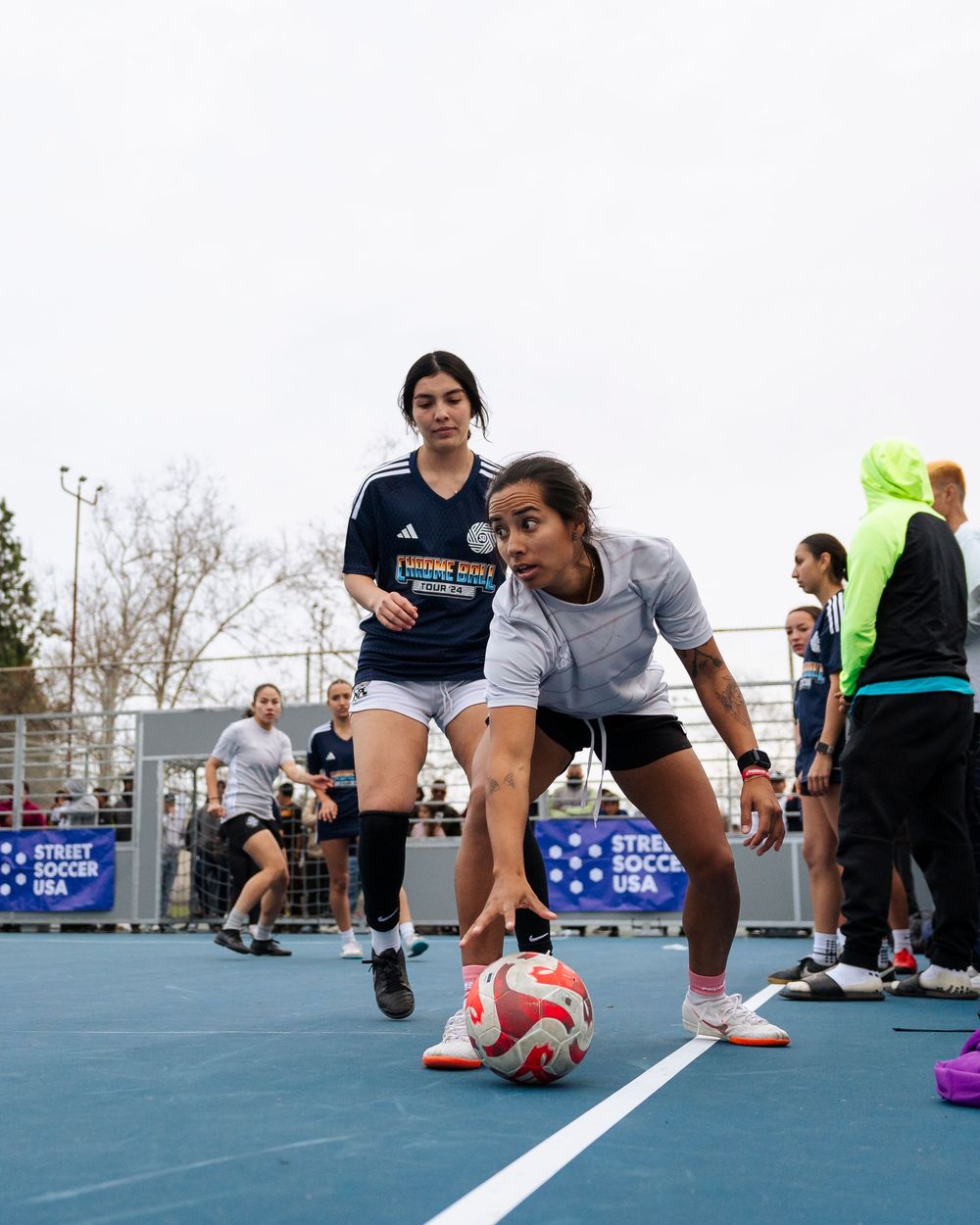 Two female soccer players stand over a ball, one player has her hand on the ball getting ready to pass it in play.