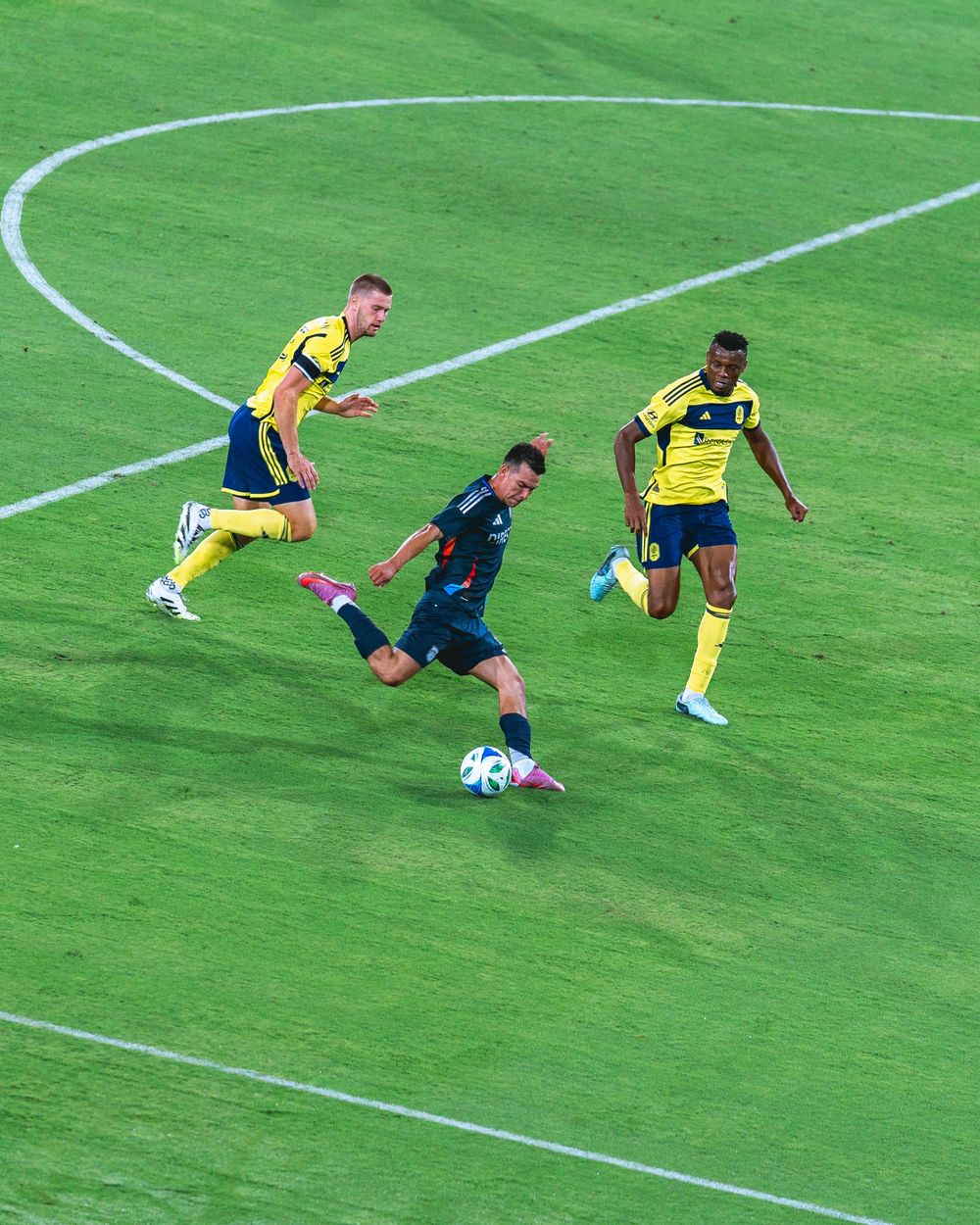 Three soccer players surround each other on the field. One of them shoots the ball towards the goal.